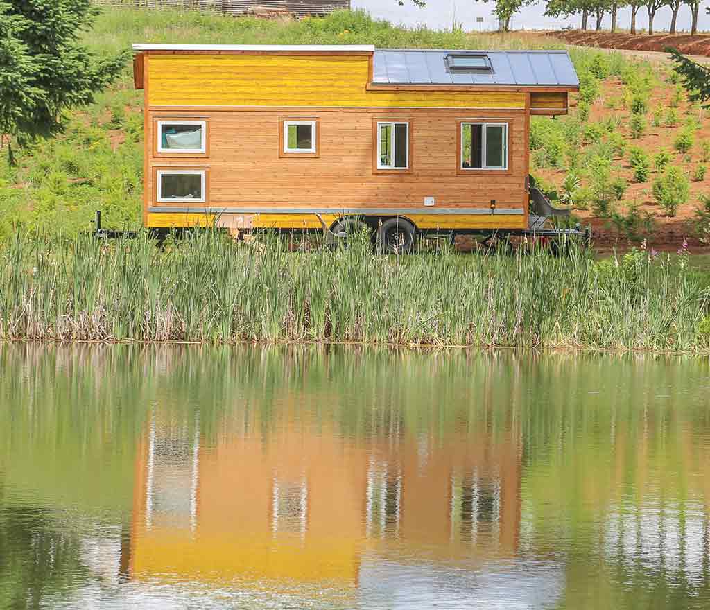 The Beachy Bohemian custom tiny home reflecting its beautiful wood and yellow outside in a lake